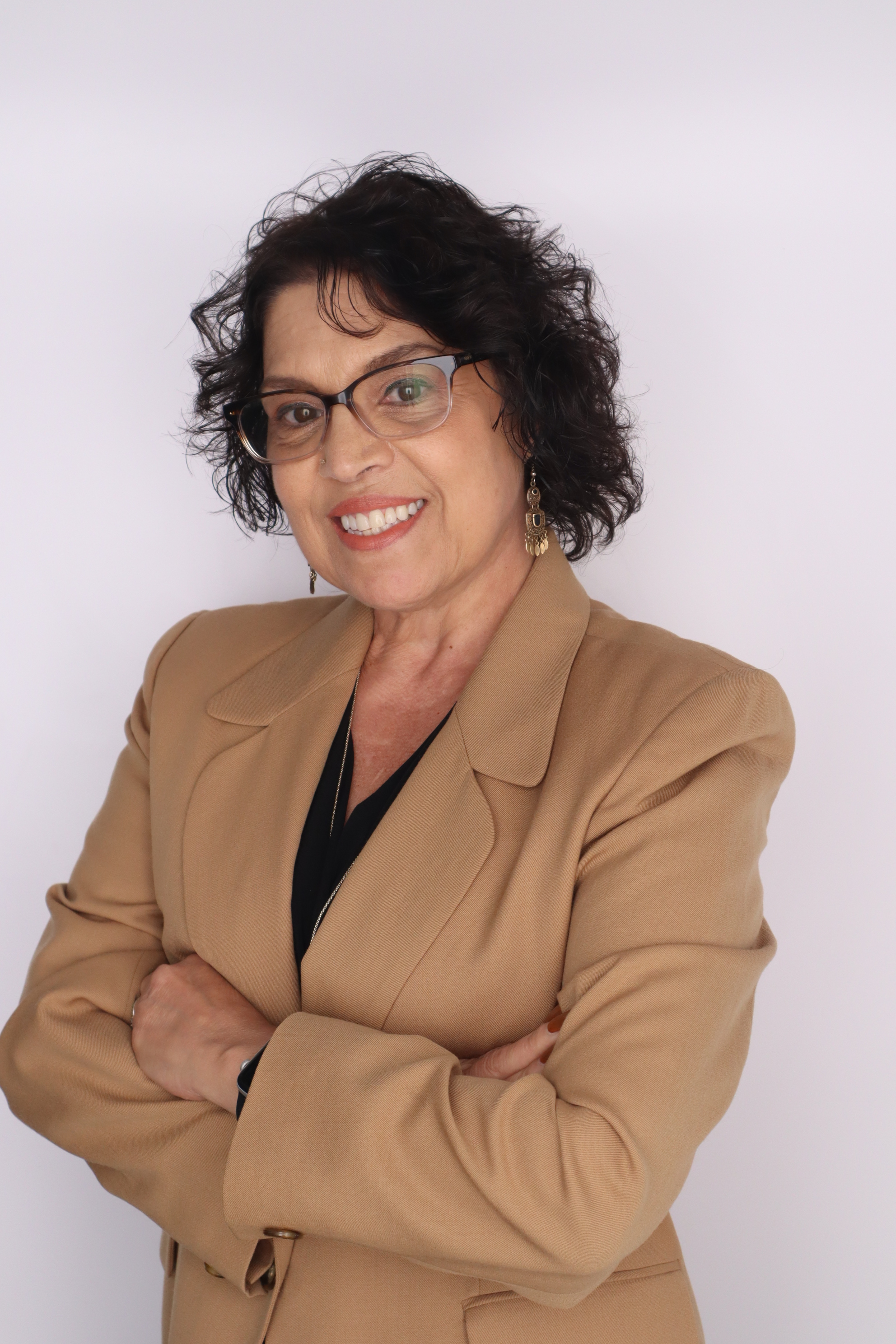 Woman smiling with brown feathered shoulder length hair. Is wearing a tan blazer with a black shirt